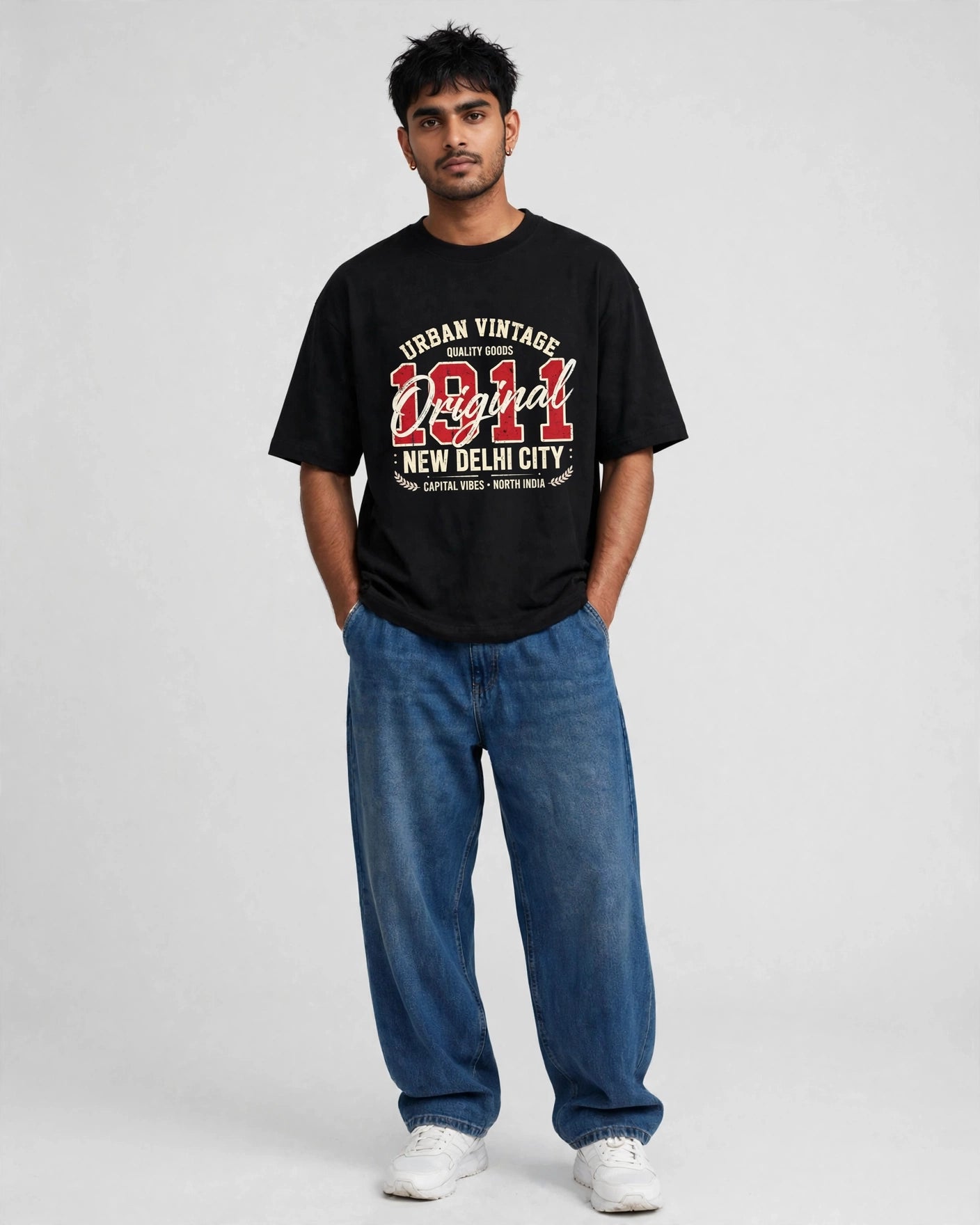 A man modeling a black graphic t-shirt with 'Urban Vintage Original New Delhi City' printed in red and white, paired with blue jeans and white sneakers, against a light gray studio background.