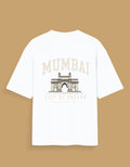 A white short-sleeve t-shirt with a graphic print of the Mumbai Gateway on the back and the text 'MUMBAI CITY OF DREAMS' in beige, displayed against a solid tan background.