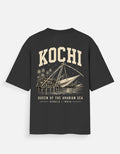 A black short-sleeve t-shirt with a vintage-style cream graphic on the back. The graphic features the word 'KOCHI' in large arched letters above an illustration of a traditional sail boat on water, with 'QUEEN OF THE ARABIAN SEA' and 'KERALA * INDIA' printed below.