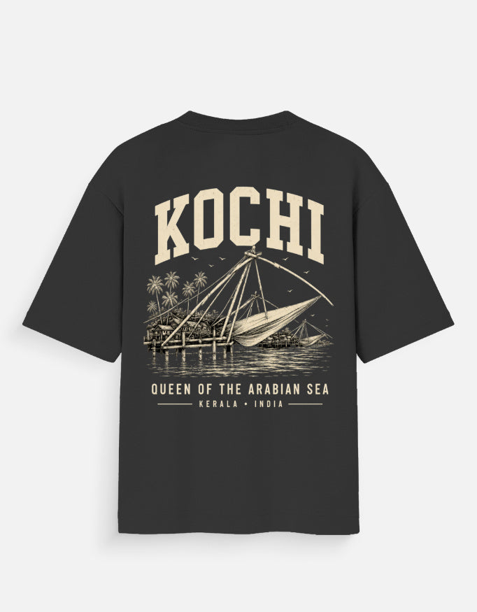 A black short-sleeve t-shirt with a vintage-style cream graphic on the back. The graphic features the word 'KOCHI' in large arched letters above an illustration of a traditional sail boat on water, with 'QUEEN OF THE ARABIAN SEA' and 'KERALA * INDIA' printed below.