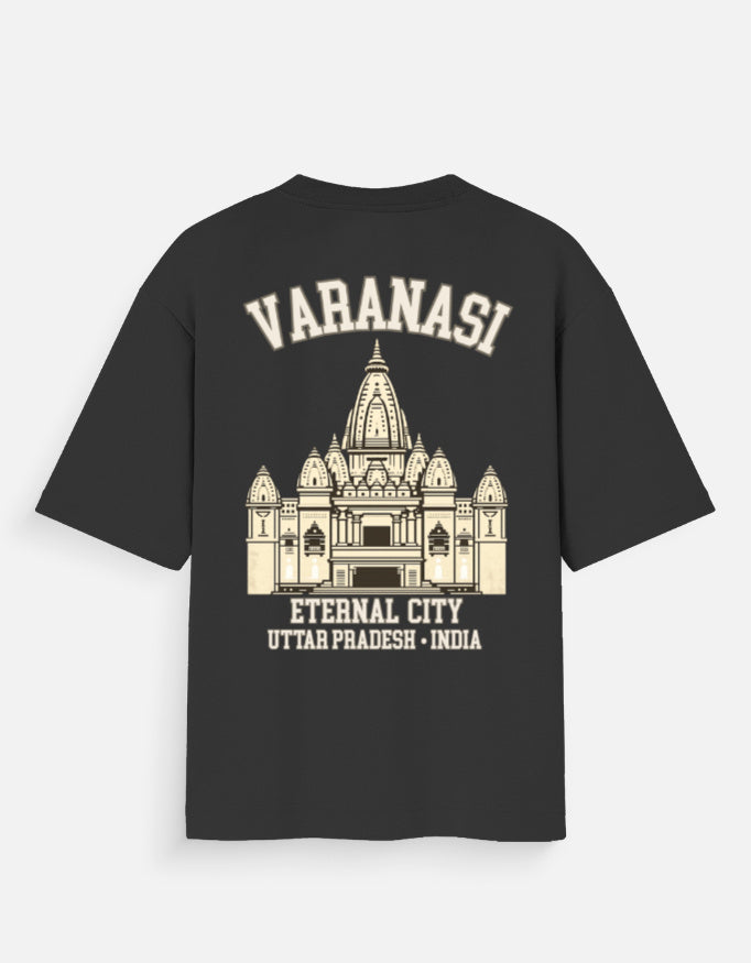 A black short-sleeve t-shirt with a cream-colored graphic print on the back. The graphic features the text 'VARANASI' in arched letters above an illustration of the Harmandir Sahib temple in Varanasi, with 'ETERNAL CITY' and 'UTTAR PRADESH - INDIA' printed below.