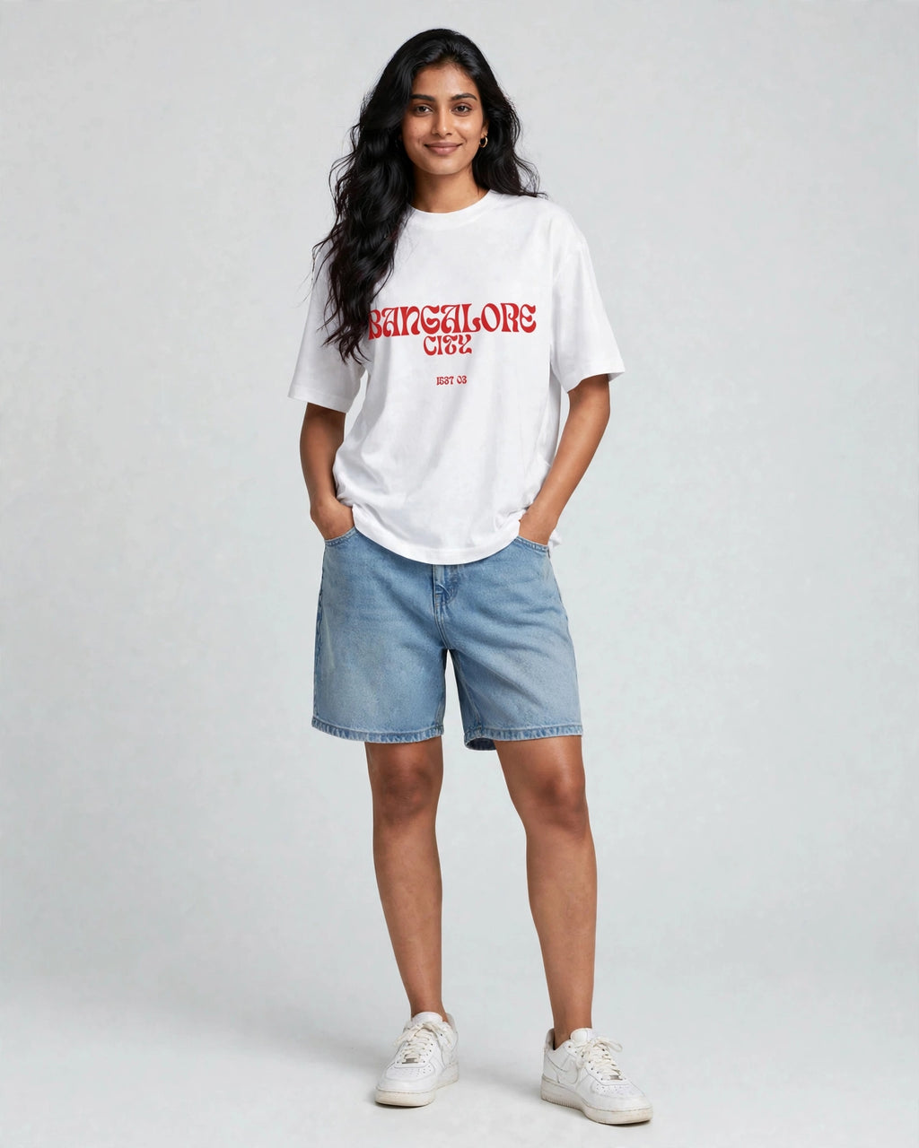 A woman modeling a white t-shirt with red 'Barcelonés' text, paired with light blue denim shorts and white sneakers, against a light gray studio background.