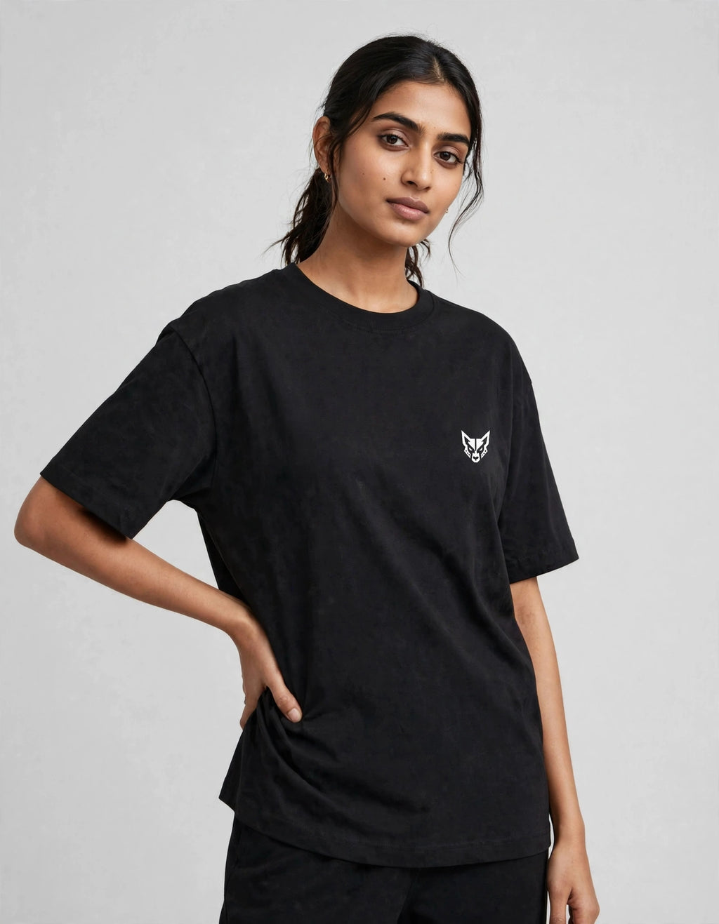 A woman modeling a black short-sleeve crewneck t-shirt with a small white graphic logo on the left chest, set against a plain light gray background.