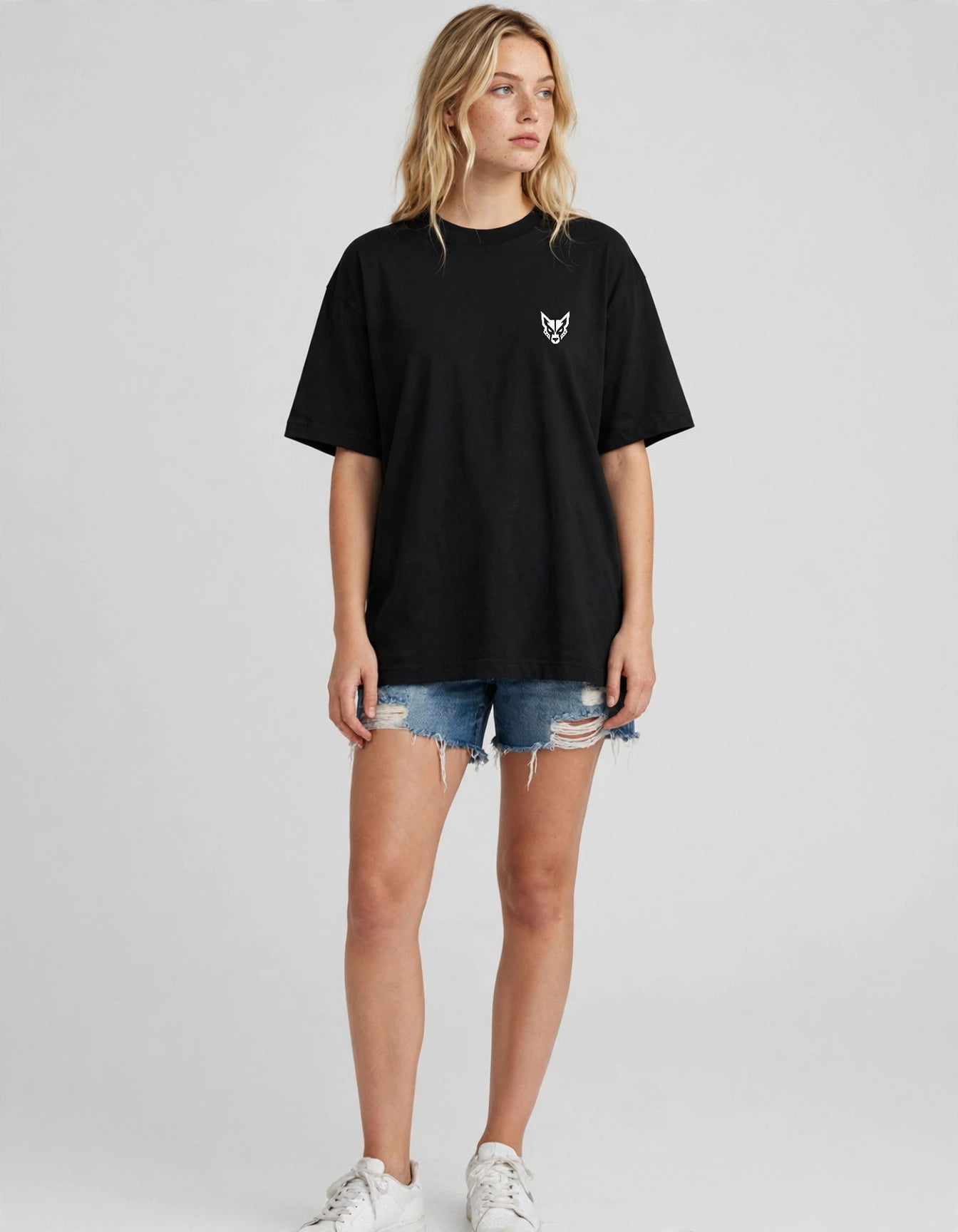 A woman modeling an oversized black short-sleeve t-shirt with a small white embroidered fox logo on the left chest, paired with distressed denim shorts and white sneakers, against a light gray studio background.