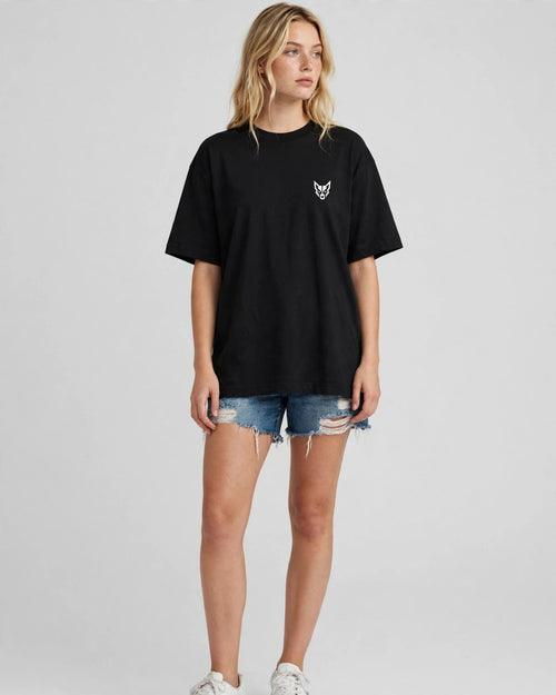 A woman modeling an oversized black short-sleeve t-shirt with a small white embroidered fox logo on the left chest, paired with distressed denim shorts and white sneakers, against a light gray studio background.