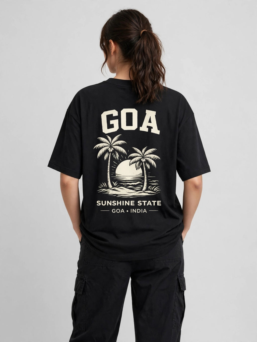 A woman viewed from behind wearing a black oversized T-shirt with a large cream graphic on the back featuring the text 'GOA', palm trees, a sunset, and 'SUNSHINE STATE GOA INDIA'.