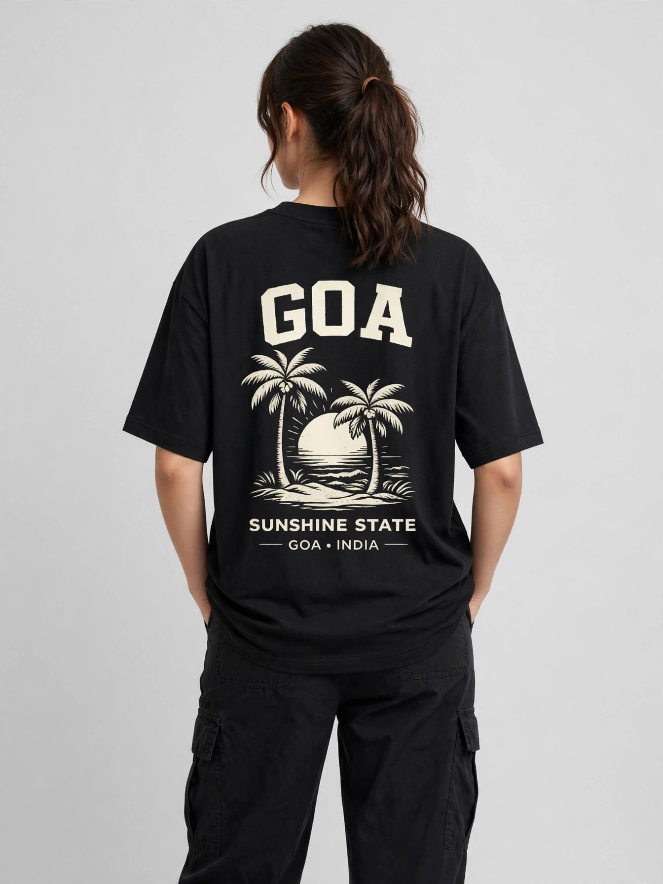A woman viewed from behind wearing a black oversized T-shirt with a large cream graphic on the back featuring the text 'GOA', palm trees, a sunset, and 'SUNSHINE STATE GOA INDIA'.