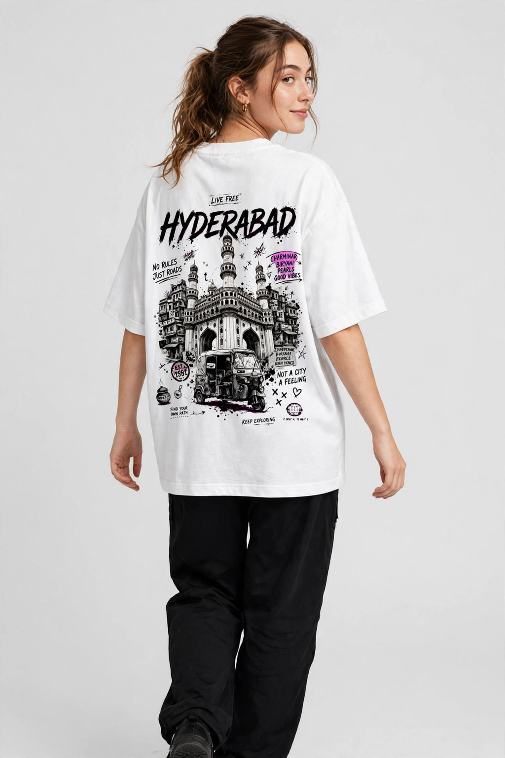 A woman modeling an oversized white graphic T-shirt from the back, featuring a black and white illustration of the Aramaq Bridge in Hyderabad with the text 'HYDERABAD' and 'Live Your Hyderabad'. She is wearing black sweatpants against a light gray studio background.