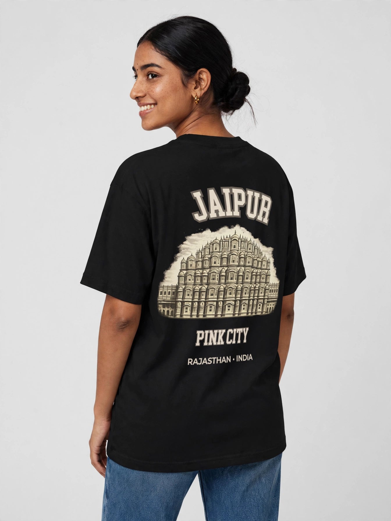 A woman shown from the back wearing a black short-sleeve graphic T-shirt with cream text reading 'JAIPUR' and 'PINK CITY' and a large building illustration, paired with blue jeans against a light gray studio background.