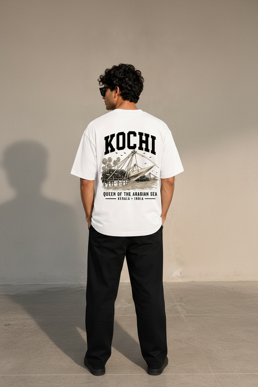 A man wearing a white graphic t-shirt with a large black print on the back reading 'KOCHI QUEEN OF THE ARABIAN SEA', paired with black pants, shown from behind against a neutral beige background.