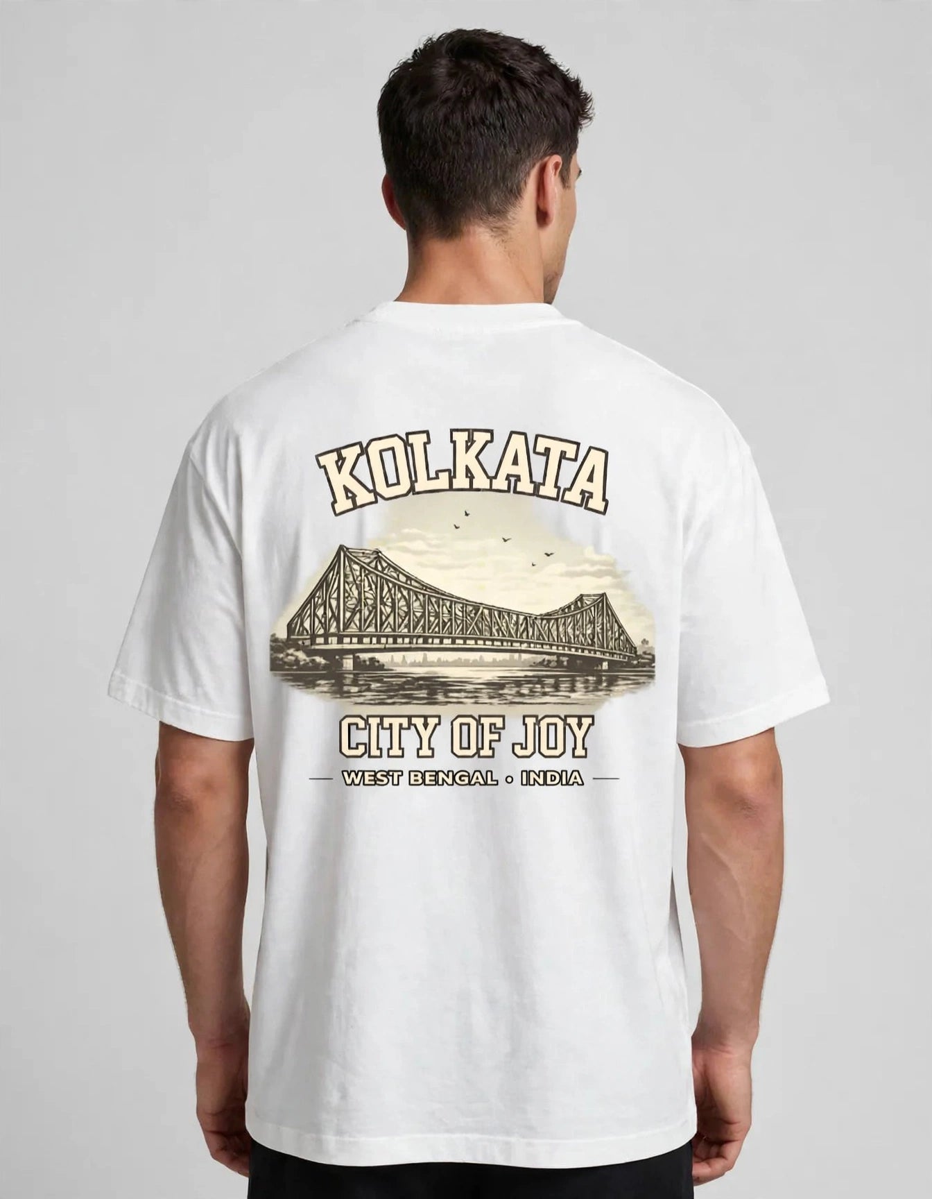 A man viewed from the back wearing a white short-sleeve T-shirt with a large vintage-style graphic on the back. The graphic features the word 'KOLKATA' above an illustration of the Kolkata bridge, with 'CITY OF JOY' and 'WEST DENSEST • BENGAL' below, in beige and brown tones.