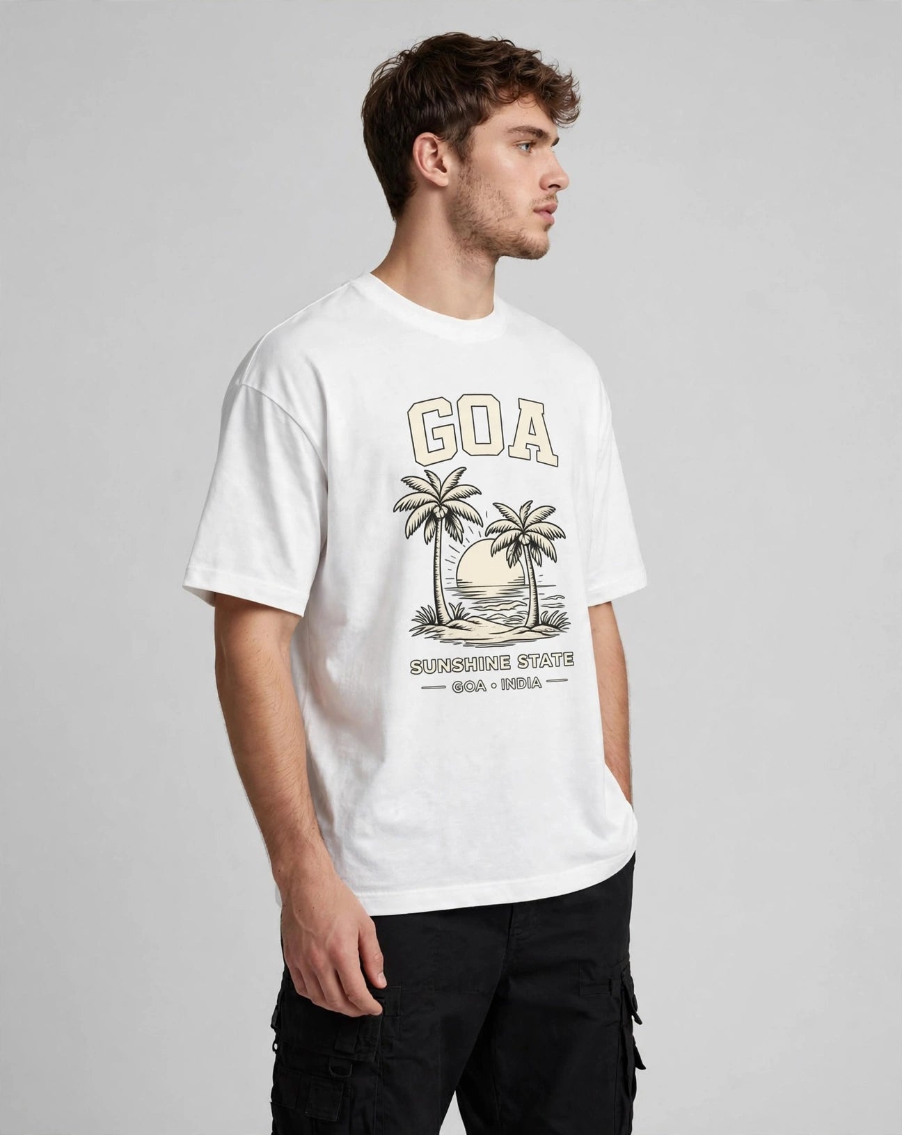 A man wearing a white short-sleeve graphic t-shirt with a vintage-style 'GOA' tropical beach print, paired with black cargo pants, photographed against a neutral gray studio background.