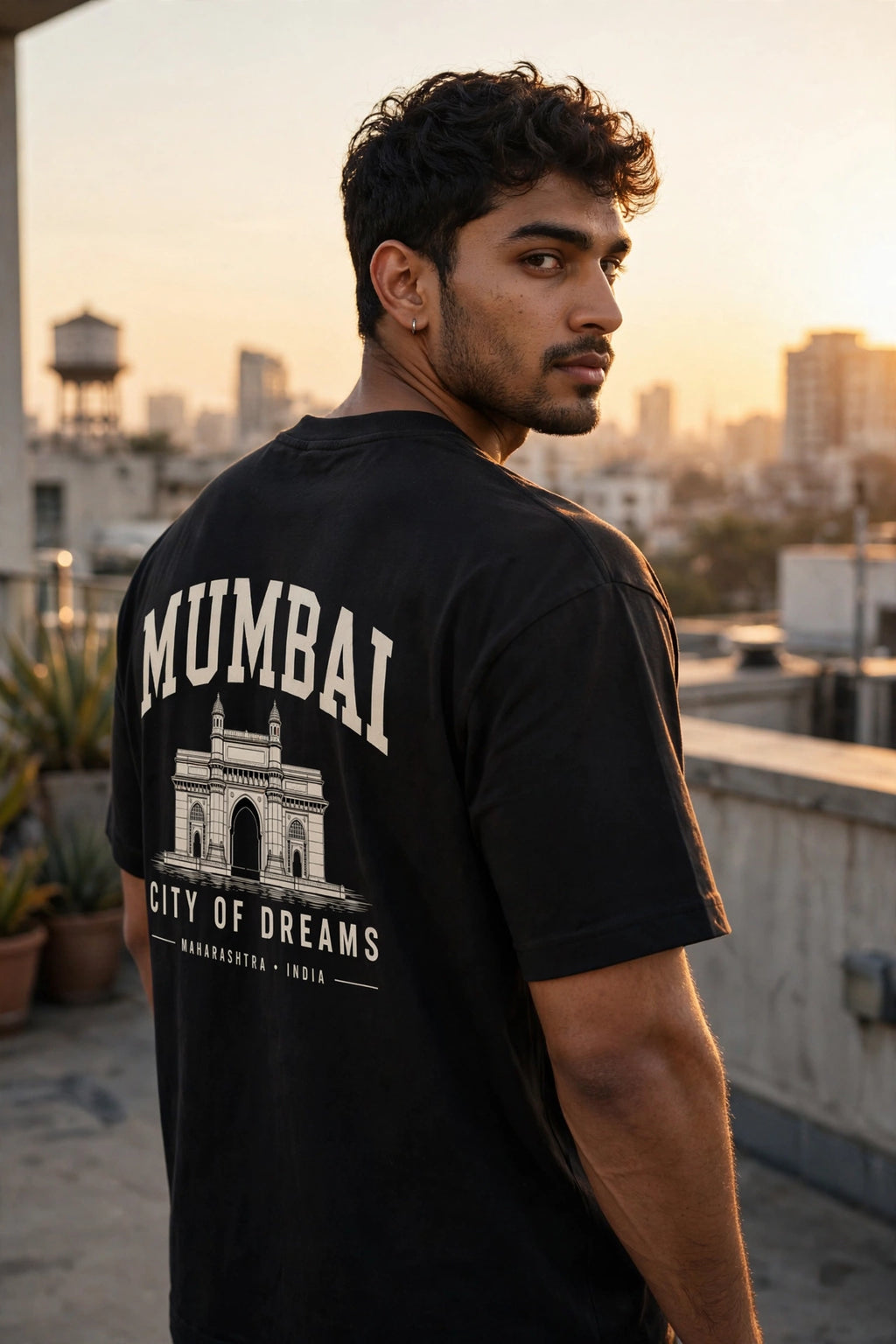A man wearing a black t-shirt with a large white graphic print on the back featuring the text 'MUMBAI' and 'CITY OF DREAMS' with an illustration of the Gateway of India, photographed on a rooftop at sunset.