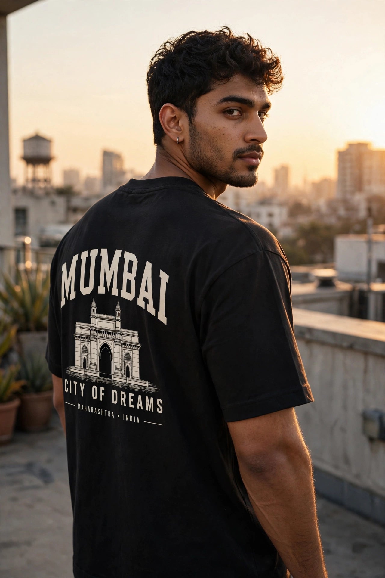 A man wearing a black t-shirt with a large white graphic print on the back featuring the text 'MUMBAI' and 'CITY OF DREAMS' with an illustration of the Gateway of India, photographed on a rooftop at sunset.