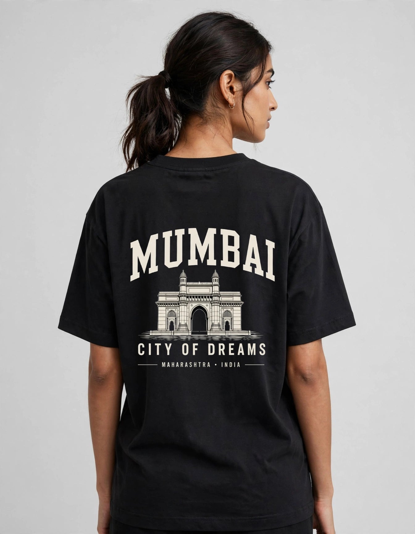 A woman shown from the back wearing a black t-shirt with a large cream-colored graphic print of Mumbai's Gateway of India and the text 'MUMBAI CITY OF DREAMS'.