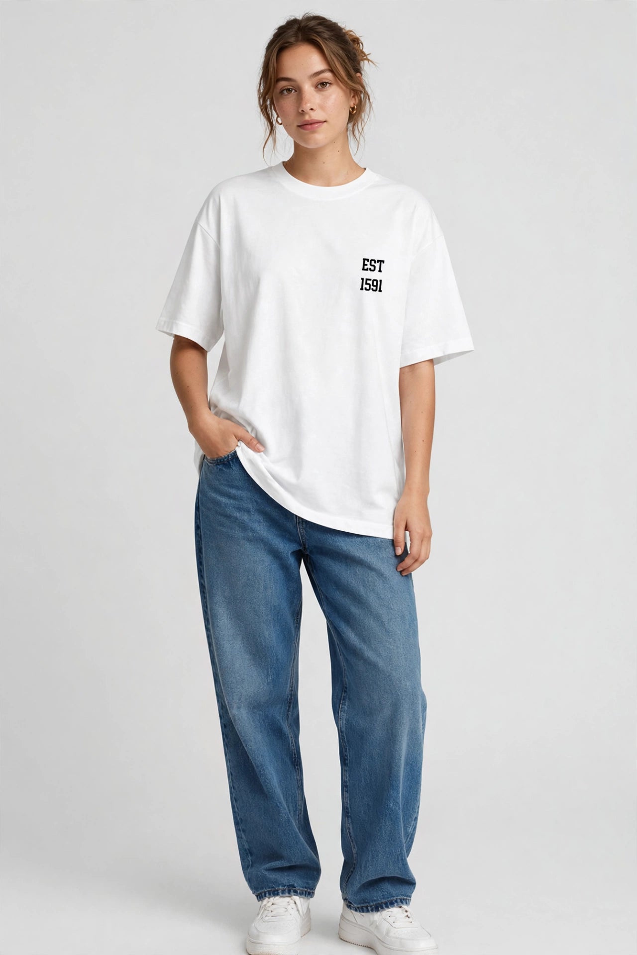 A woman modeling an oversized white t-shirt with a small black 'EST 1996' graphic on the left chest, paired with blue jeans and white sneakers, against a light gray studio background.