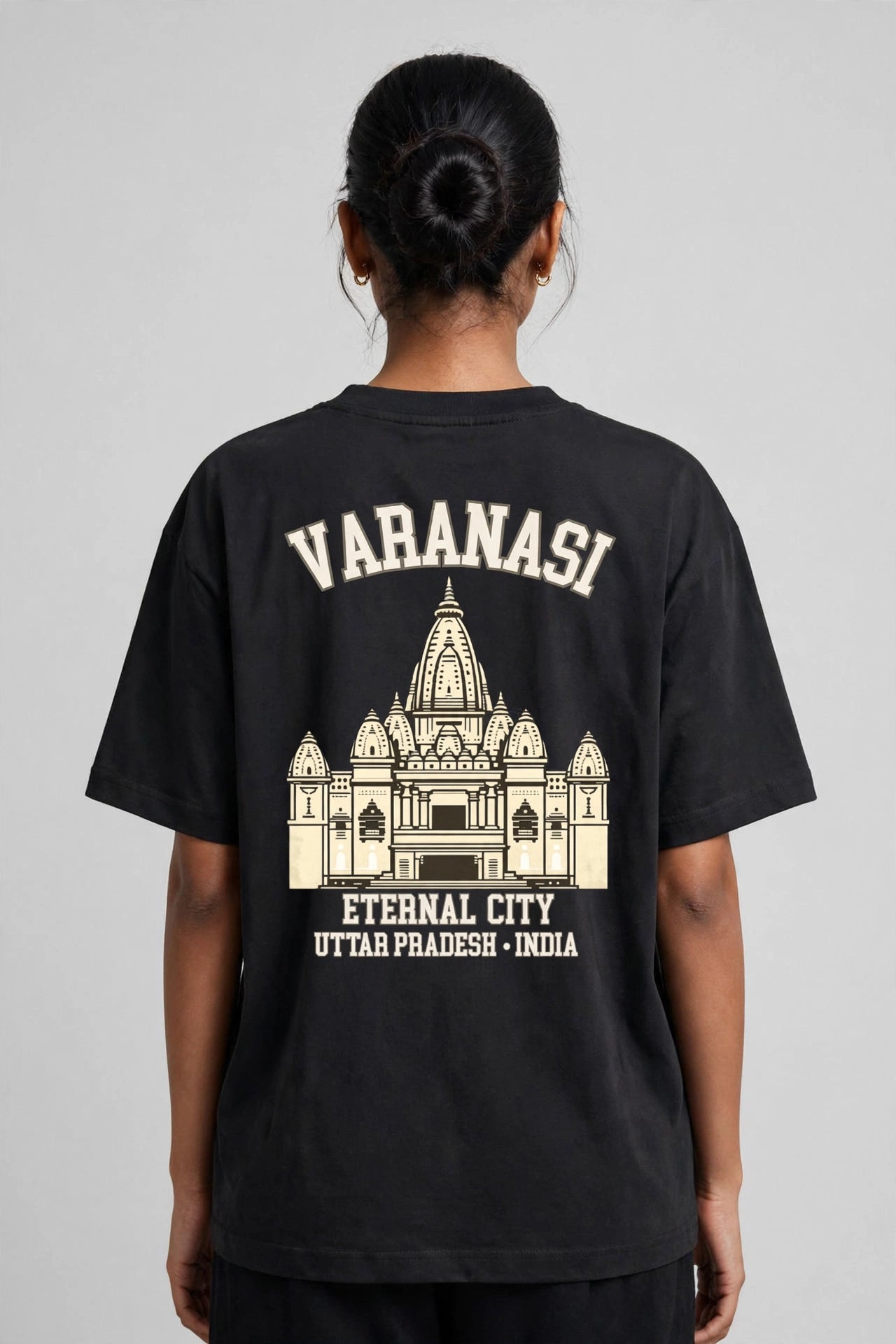 A person shown from the back wearing a black oversized T-shirt with a large cream-colored graphic print featuring the text 'VARANASI', an illustration of a temple, and 'ETERNAL CITY UTTAR PRADESH - INDIA'.