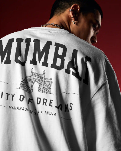 A close-up of a person from behind wearing a white oversized T-shirt with large black 'MUMBAI' lettering and a black architectural graphic on the back, against a dark red studio background.