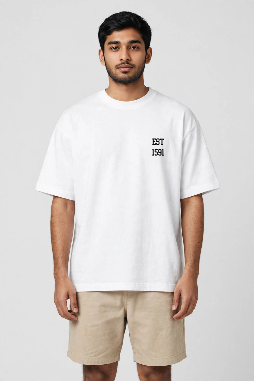 A man wearing a white oversized crewneck t-shirt with 'EST 1591' printed in black on the left chest, paired with beige shorts, against a light gray studio background.
