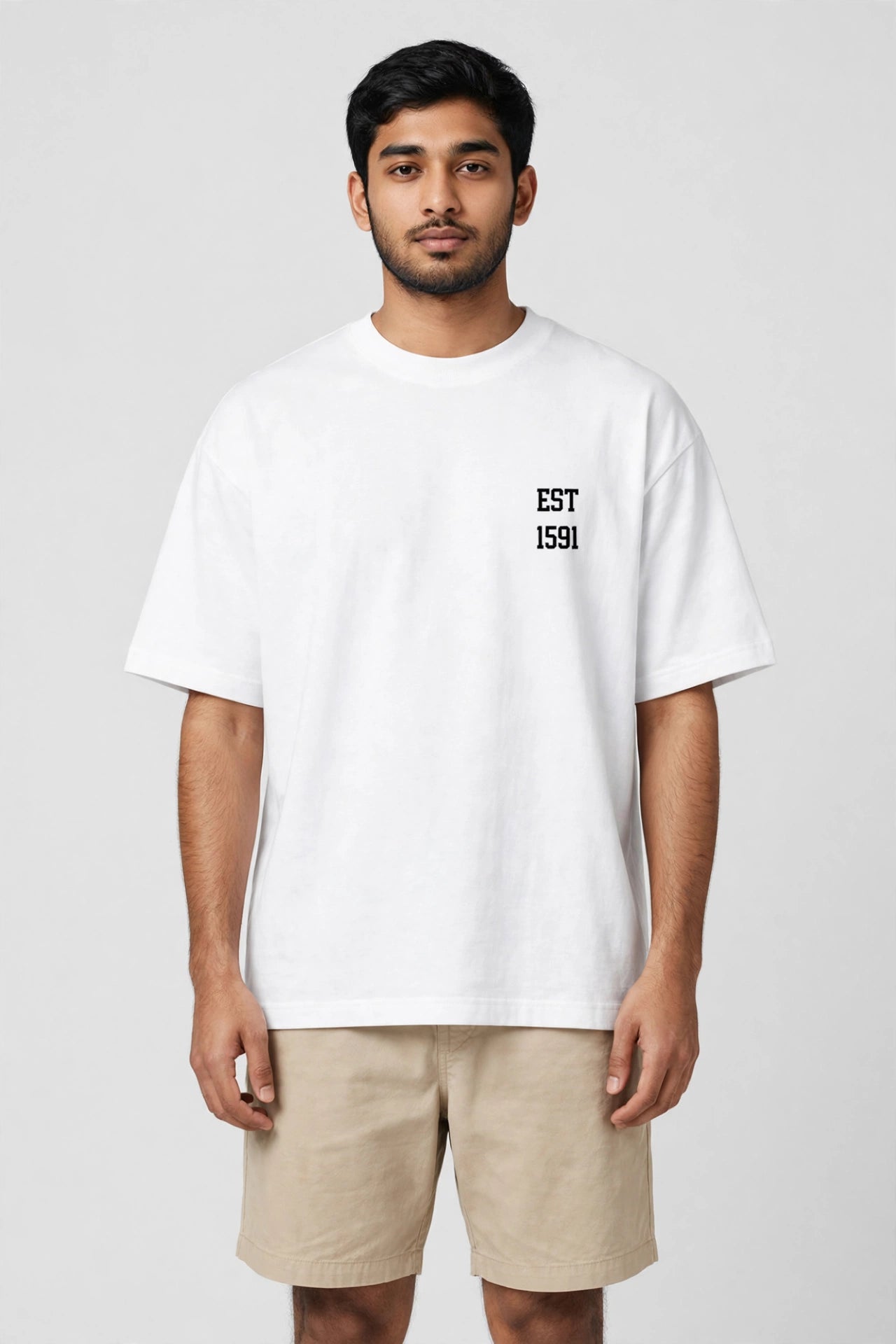 A man wearing a white oversized crewneck t-shirt with 'EST 1591' printed in black on the left chest, paired with beige shorts, against a light gray studio background.