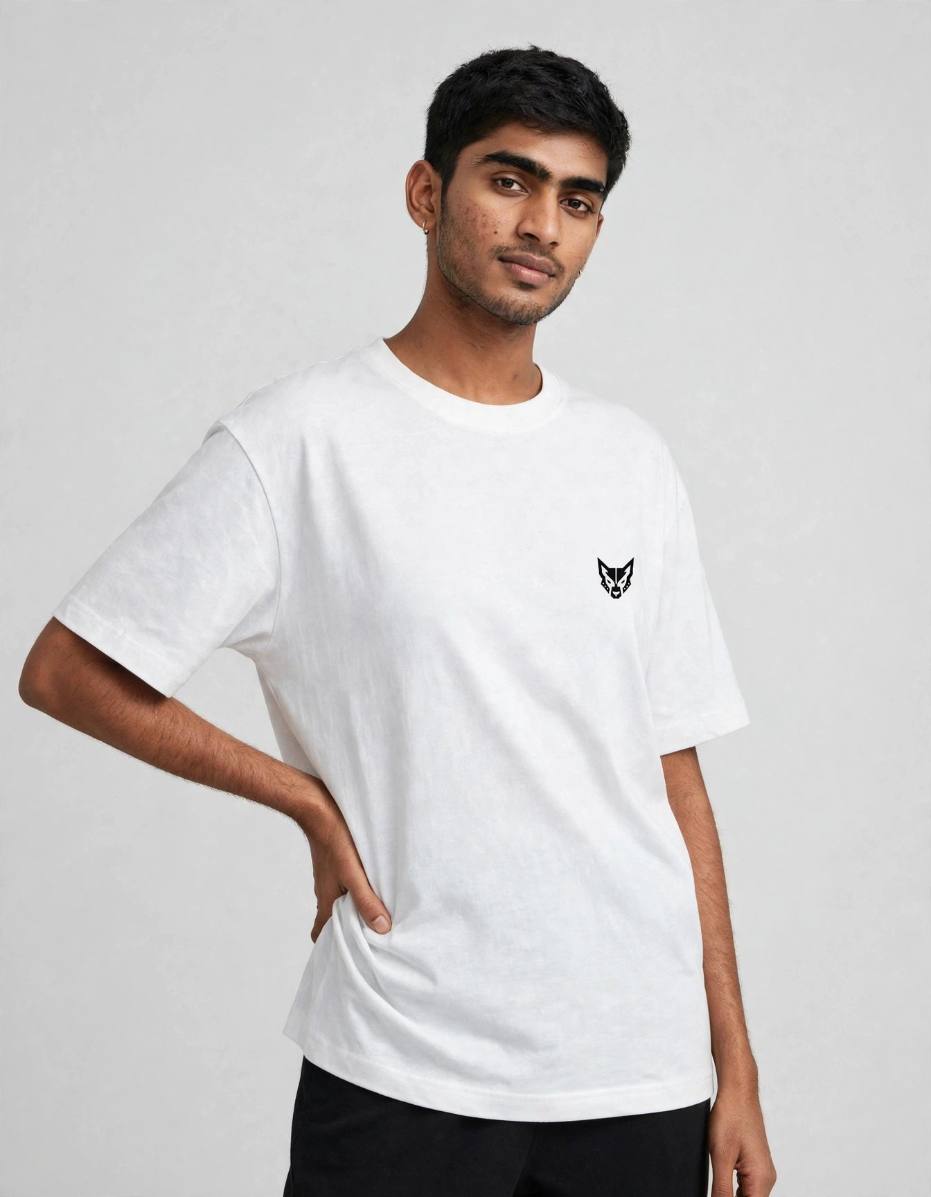 A man wearing a white short-sleeve crewneck t-shirt with a small black embroidered fox head logo on the left chest, paired with black pants, against a light gray studio background.