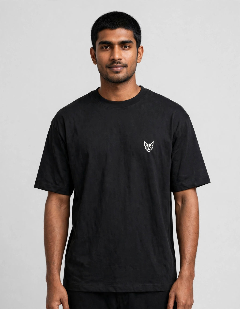 A man wearing a black short-sleeve crewneck t-shirt with a small white embroidered fox head logo on the left chest, photographed against a neutral light gray background.