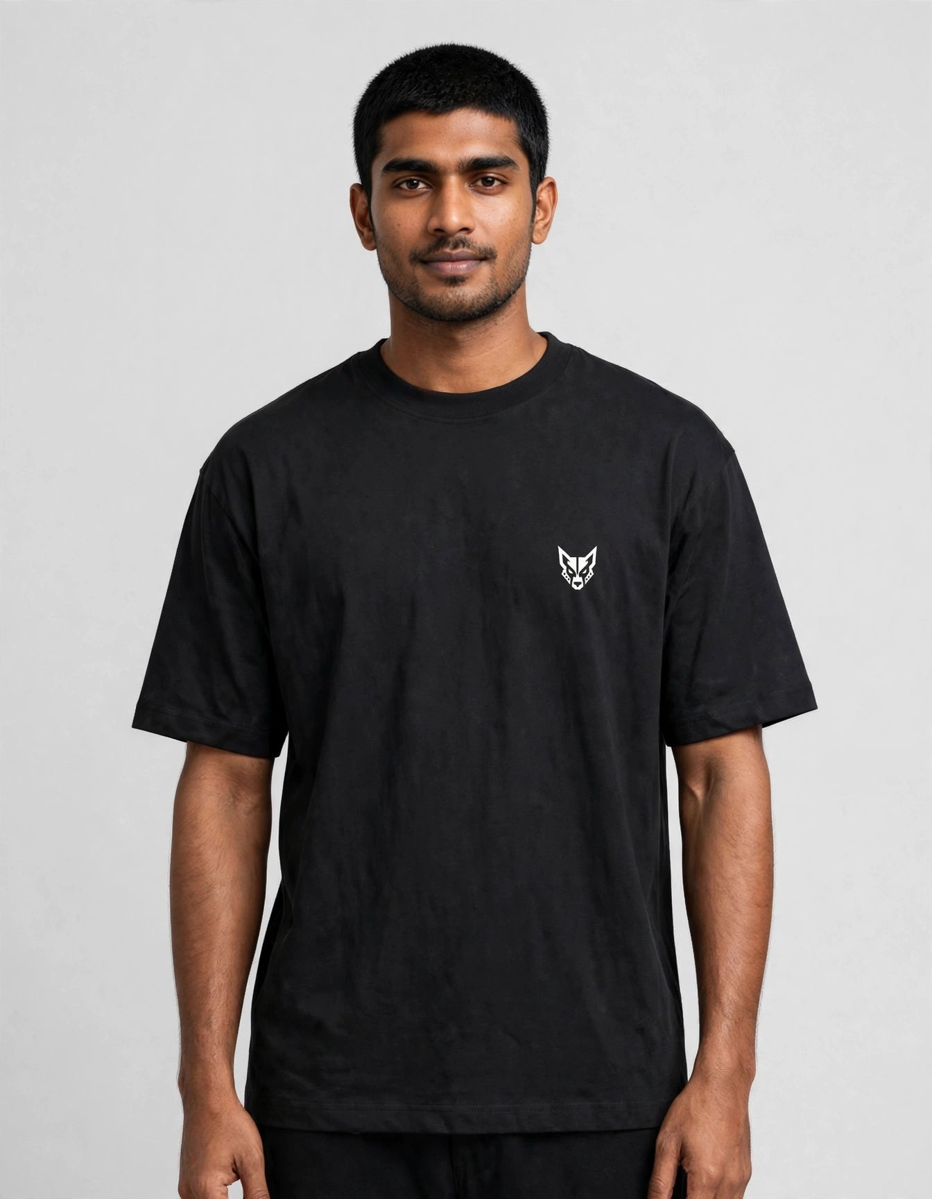 A man wearing a black short-sleeve crewneck t-shirt with a small white embroidered fox head logo on the left chest, photographed against a neutral light gray background.