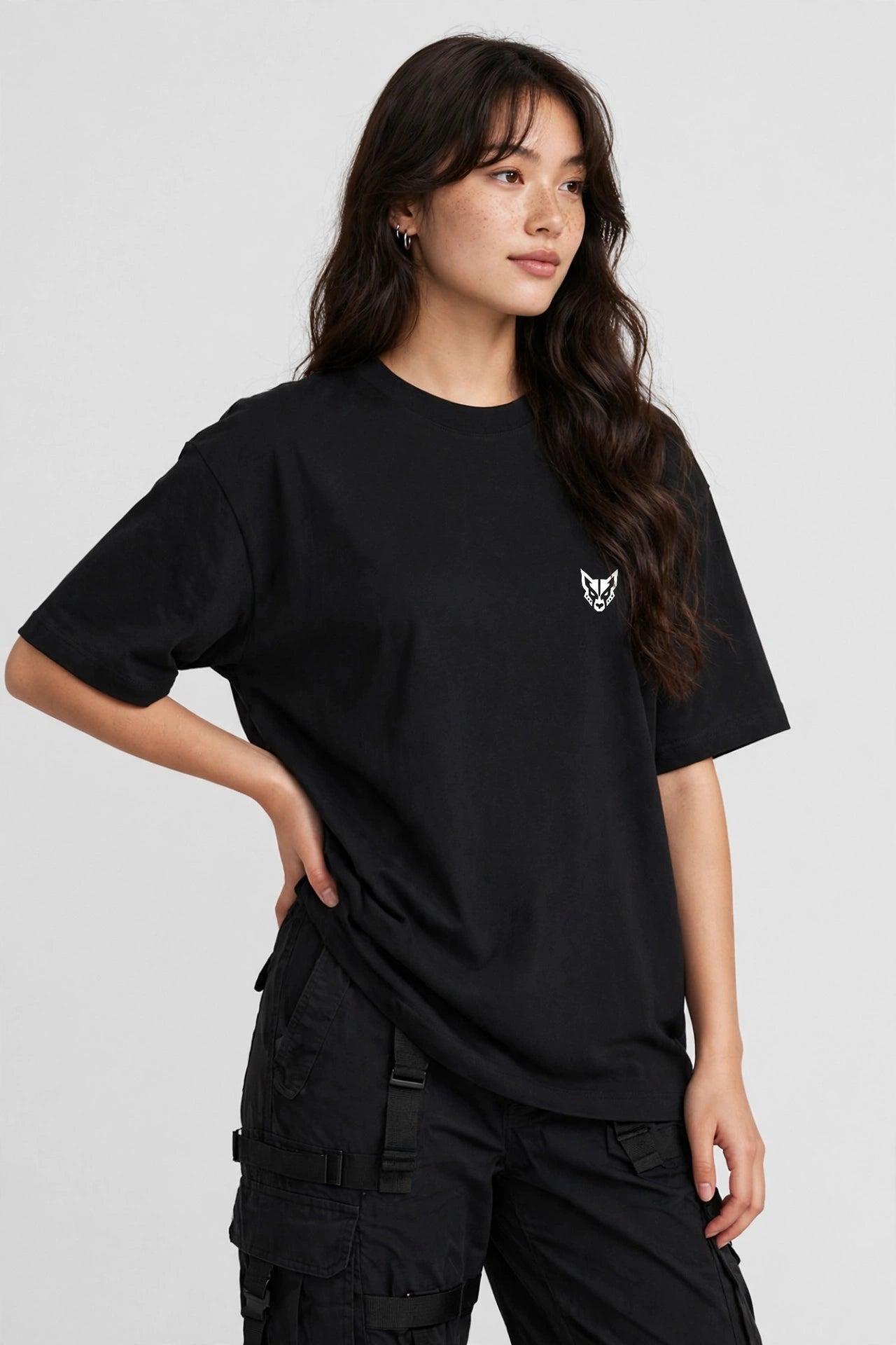 A woman wearing an oversized black short-sleeve t-shirt with a small white embroidered fox head logo on the left chest, paired with black cargo pants, against a light gray studio background.