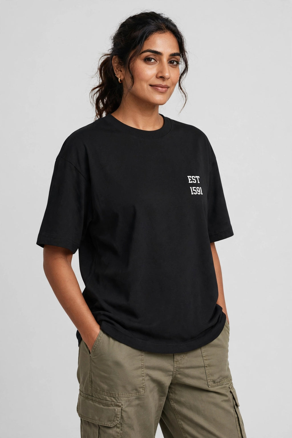 A woman wearing a black short-sleeve t-shirt with a small white 'EST. 1551' graphic on the left chest, paired with olive green cargo pants, against a light gray studio background.
