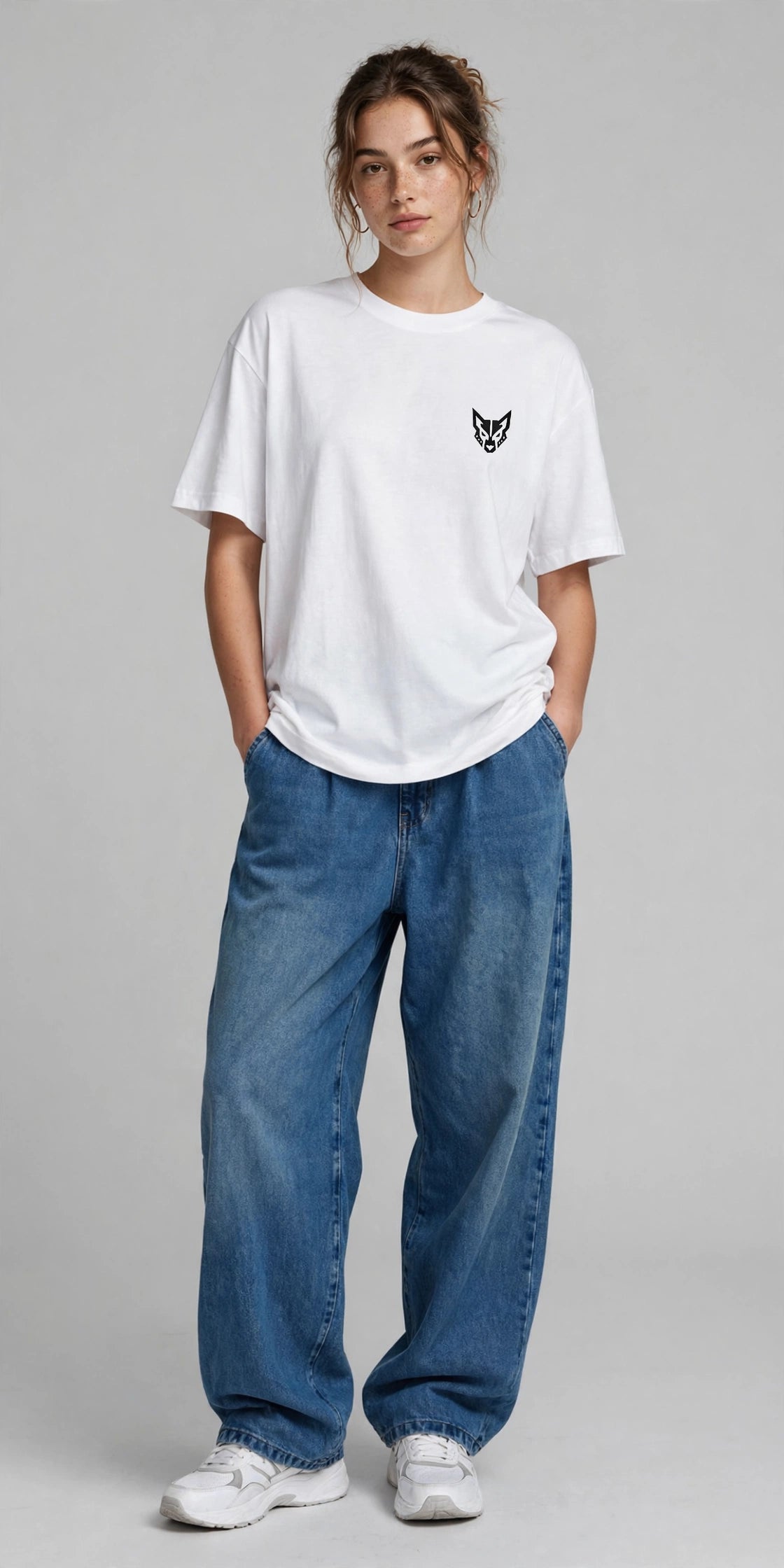 A woman modeling a white short-sleeve t-shirt with a small black chest graphic, paired with loose blue jeans and white sneakers, against a solid gray studio background.