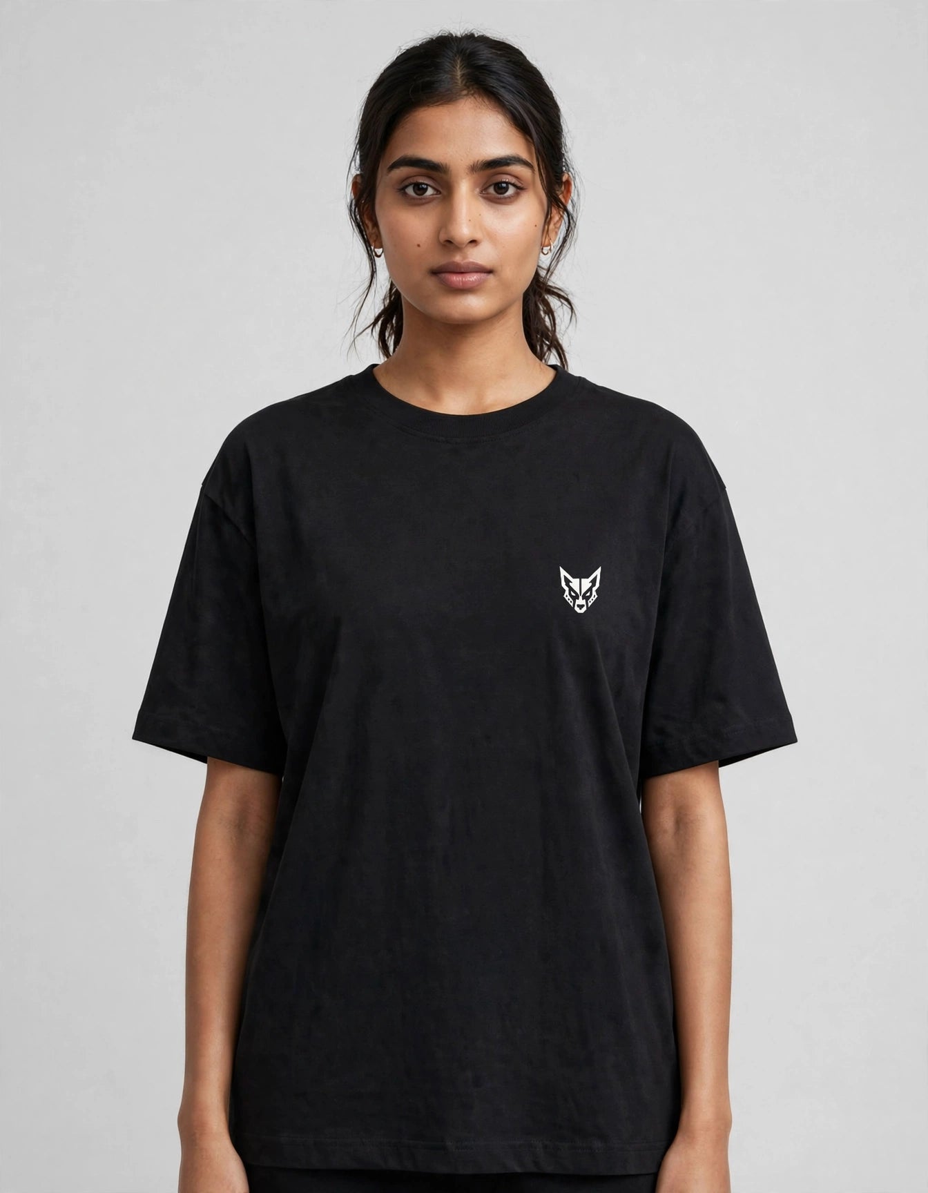 A woman wearing a black short-sleeve crewneck t-shirt with a small white embroidered fox head logo on the left chest, photographed against a neutral gray studio background.