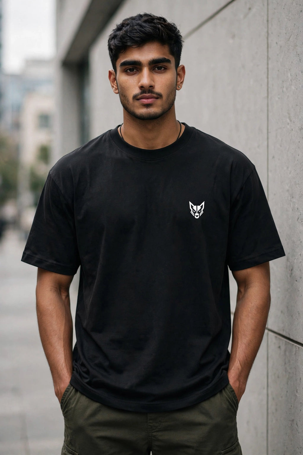 A man wearing a black crewneck t-shirt with a small white wolf head logo on the left chest, standing outdoors against a gray concrete wall.