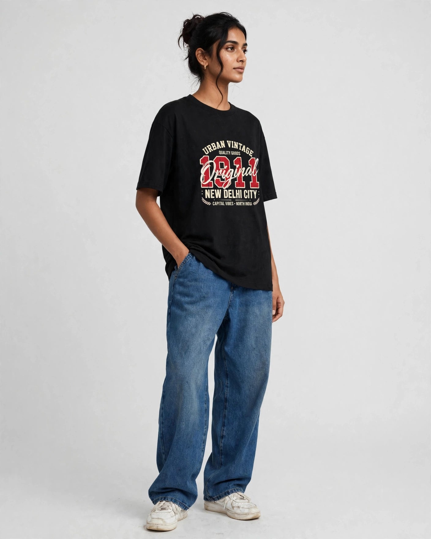 A woman modeling a black graphic t-shirt with a red and white 'New Delhi City' print, paired with blue jeans and white sneakers, against a light gray studio background.