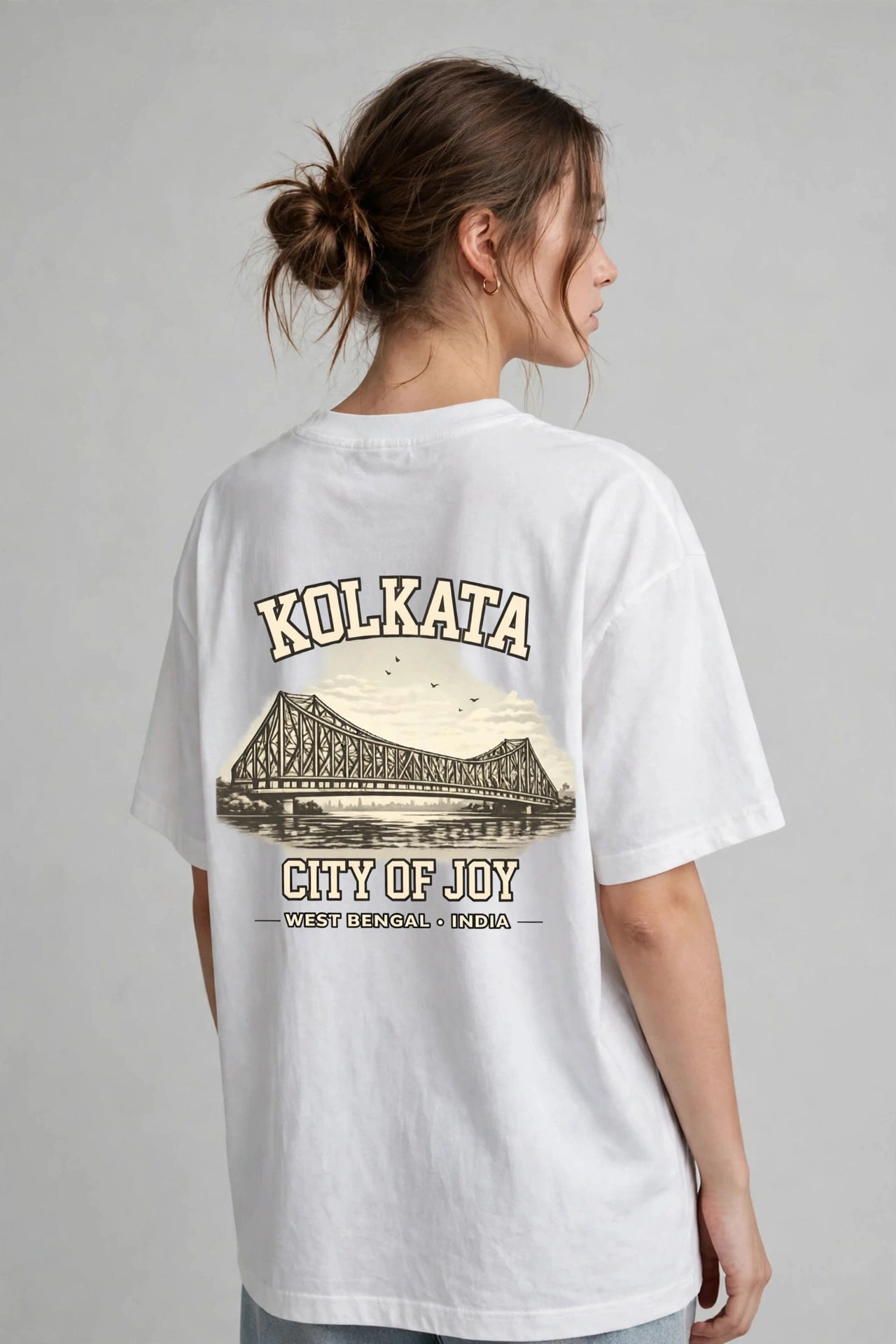 A woman shown from the back wearing an oversized white graphic T-shirt with a vintage-style Kolkata bridge print and the text 'KOLKATA' and 'CITY OF JOY'.