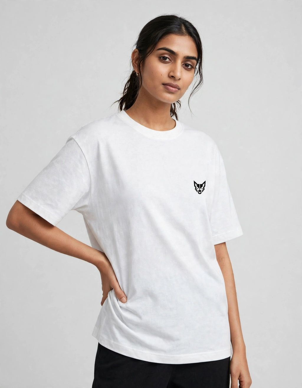 A woman modeling a white short-sleeve crewneck t-shirt with a small black fox head logo on the left chest, paired with black pants, against a light gray studio background.