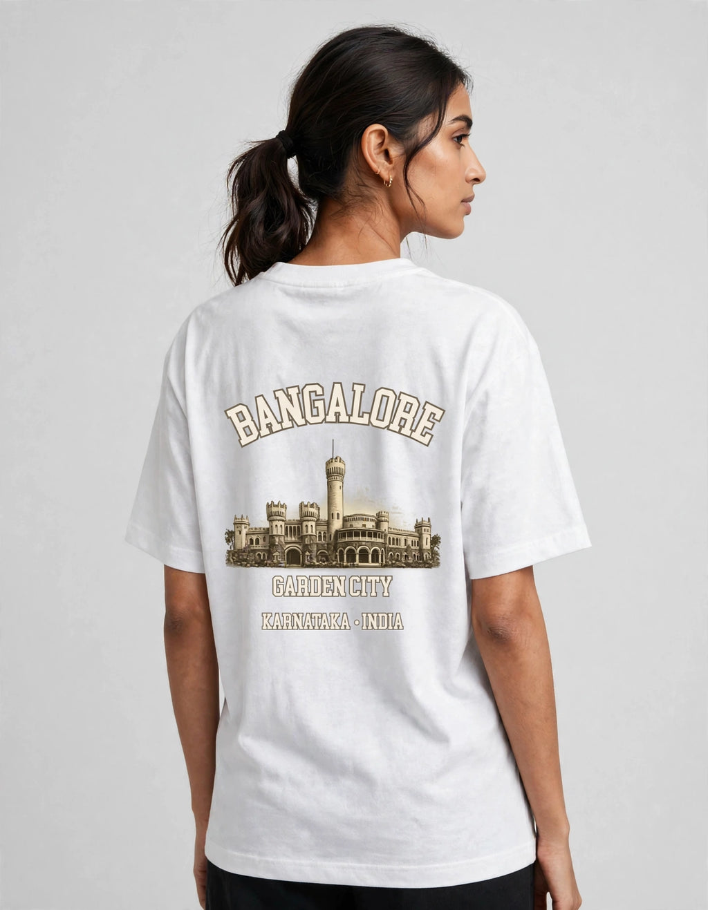 A woman shown from the back wearing a white short-sleeve T-shirt with a large beige and brown graphic print on the back featuring the word 'BANGALORE' and a cityscape illustration.