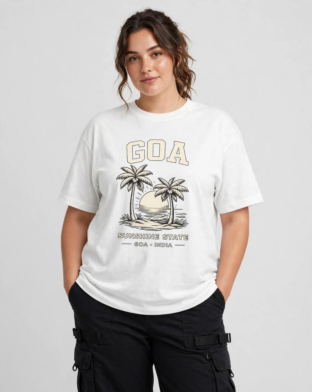 A woman modeling a white graphic t-shirt featuring a vintage-style 'GOA' beach graphic with palm trees and 'SILAPUDDH STATE GOA + INDIA' text, paired with black cargo pants against a light gray studio background.