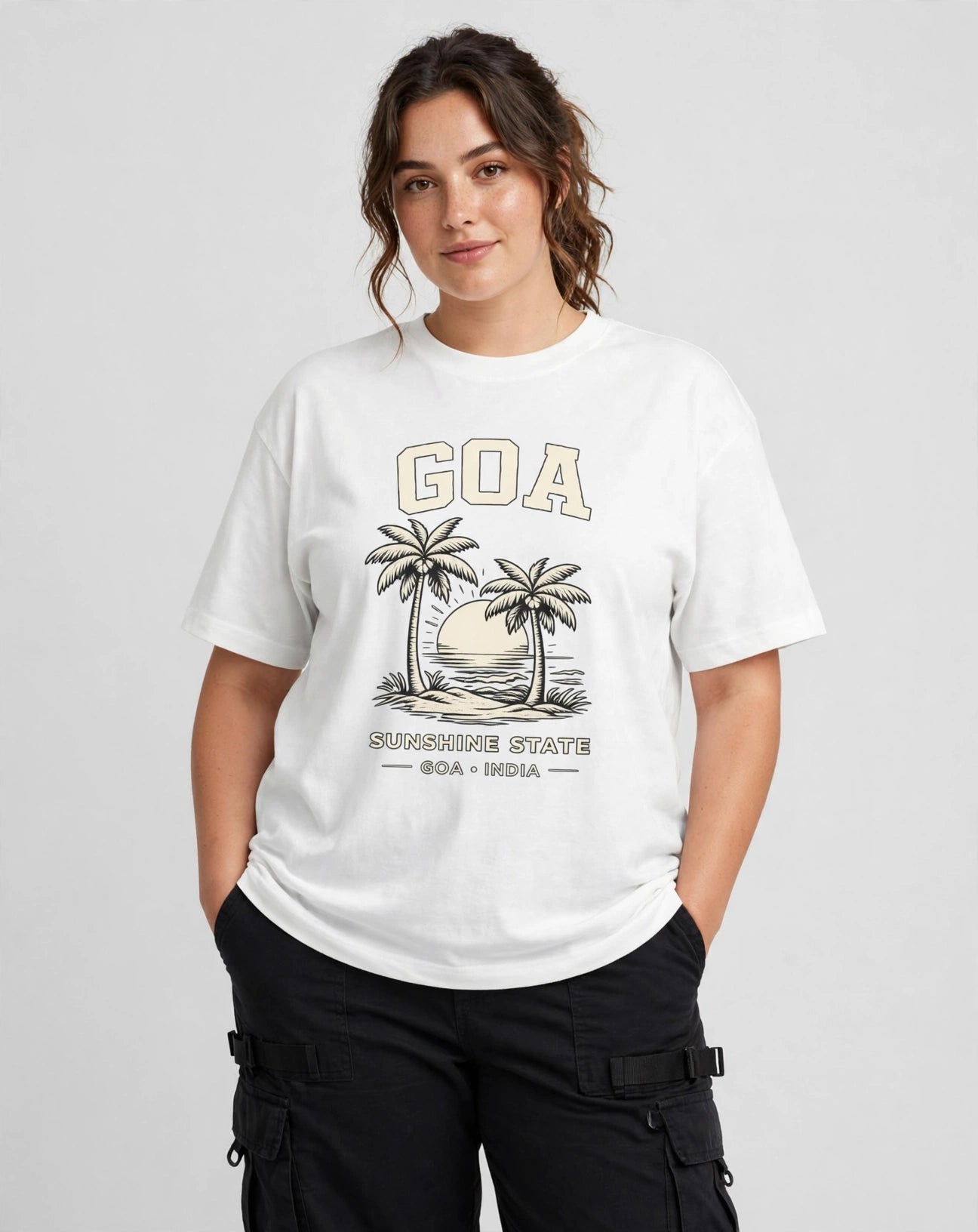 A woman modeling a white graphic t-shirt featuring a vintage-style 'GOA' beach graphic with palm trees and 'SILAPUDDH STATE GOA + INDIA' text, paired with black cargo pants against a light gray studio background.