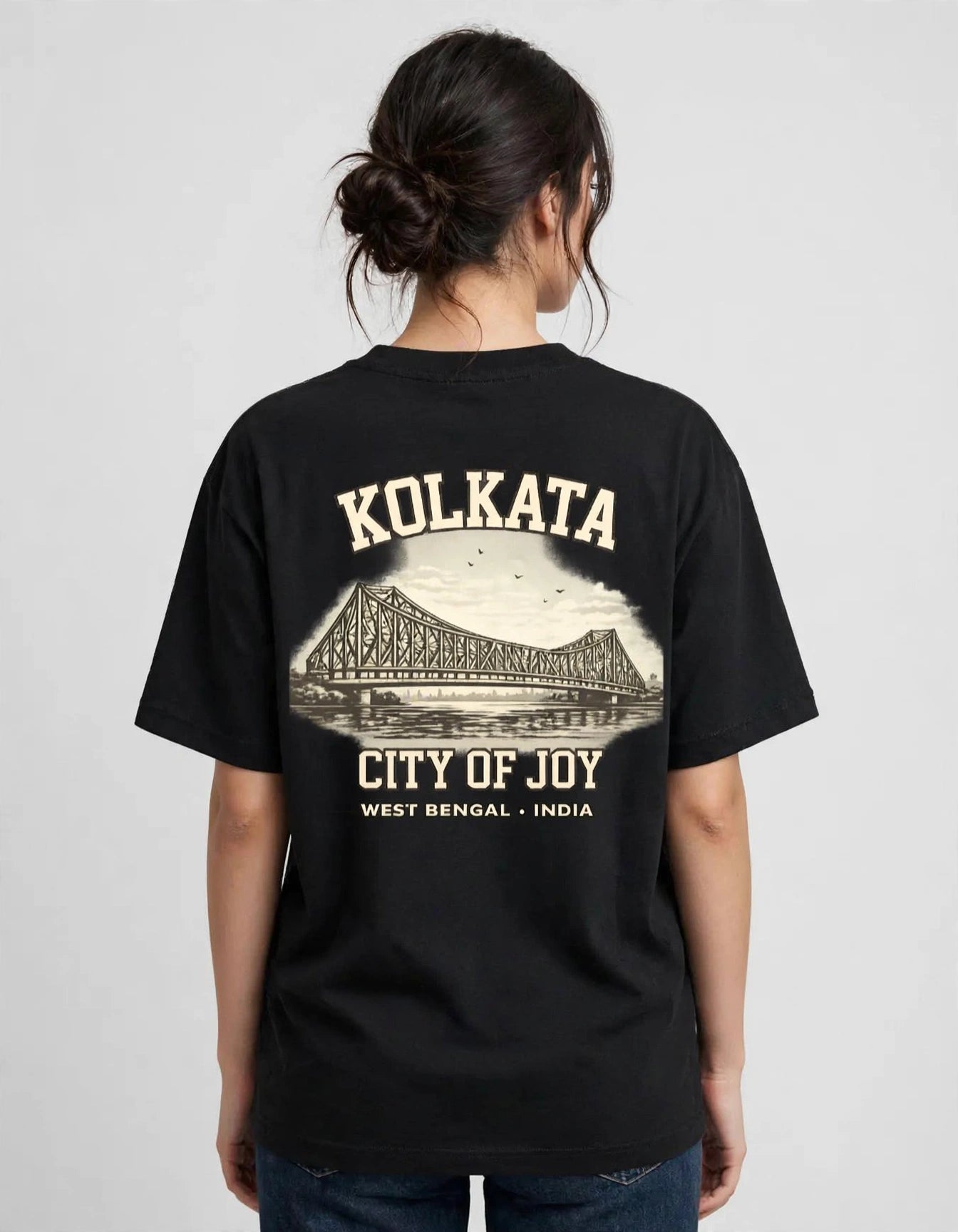 A woman viewed from the back wearing a black oversized T-shirt with a large cream-colored graphic print of the Kolkata bridge and text reading 'KOLKATA', 'CITY OF JOY', and 'WEST BENGAL - INDIA'.